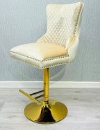 A cream colored bar stool with gold accents, featuring a tufted backrest and a lion knocker on the back.
