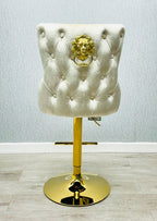 A cream colored bar stool with gold accents, featuring a tufted backrest and a lion knocker on the back.